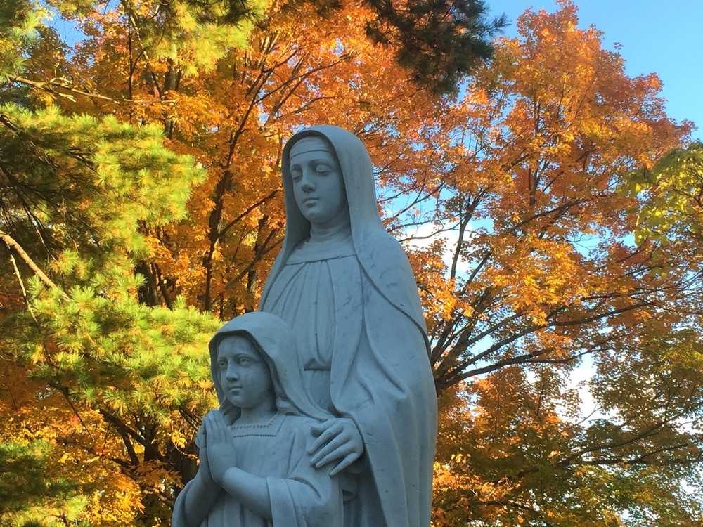 A white statue of a mother and child beneath autumn leaves on the shrine grounds.